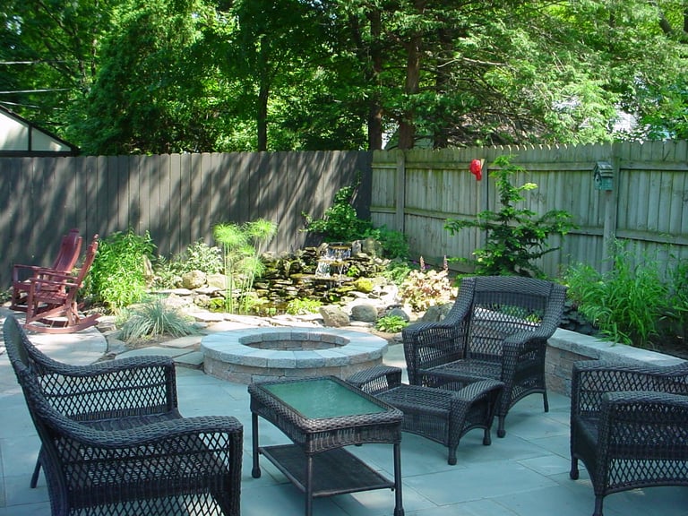 Backyard patio with firepit, wicker seating, and lush trees providing shade