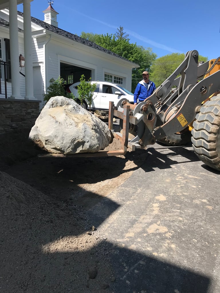 Worker operating a front-end loader to move a large boulder near a white colonial-style house on a sunny day
