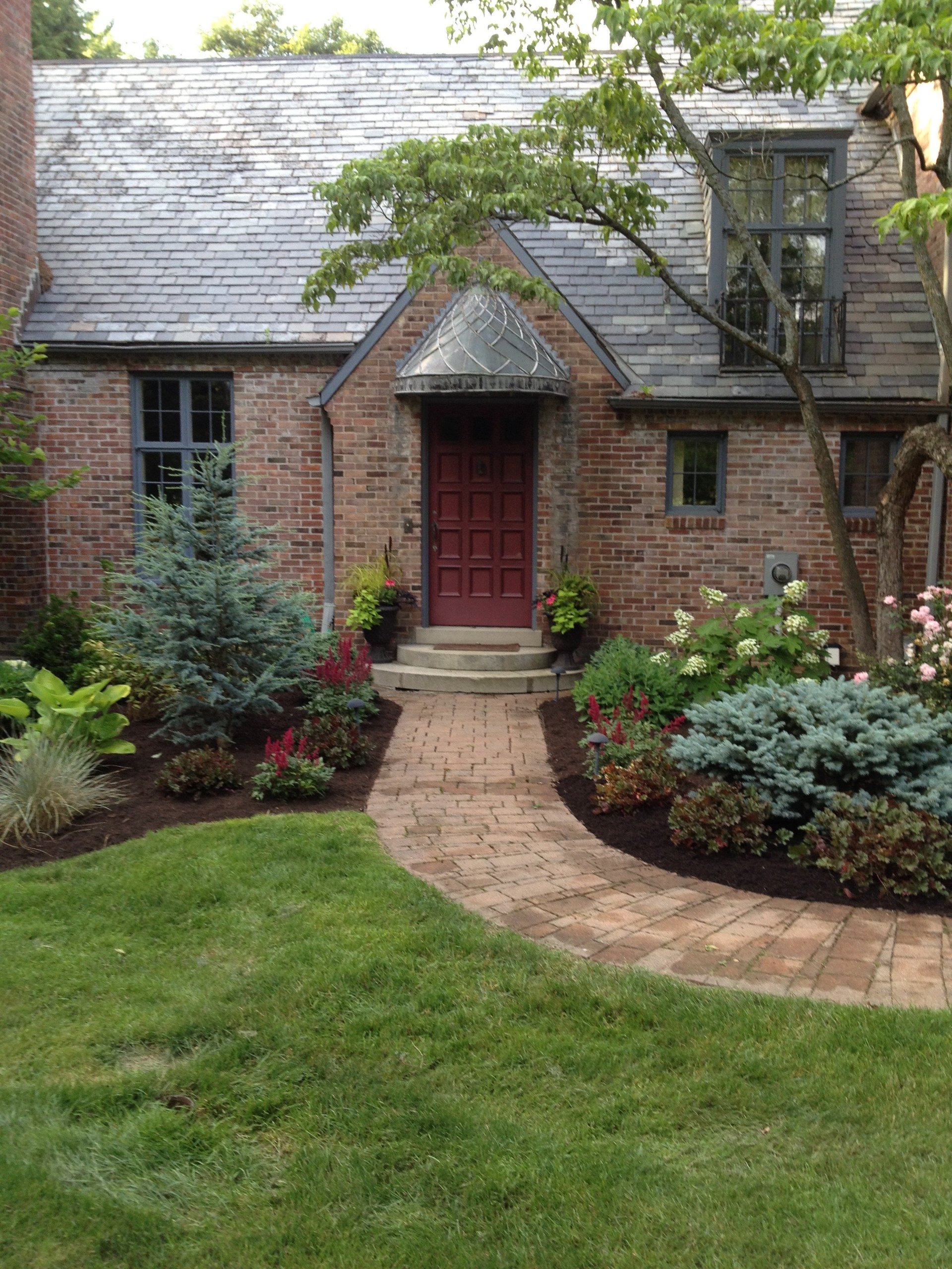 Brick residential home with red front door, slate roof, and manicured landscaping featuring shrubs and green lawn