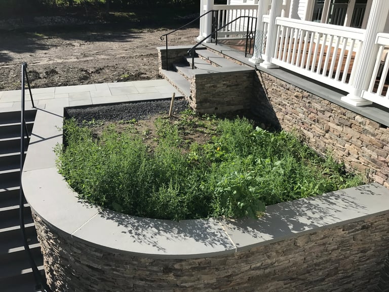 Curved stone planter bed with overgrown green plants in front of white railing on residential patio area