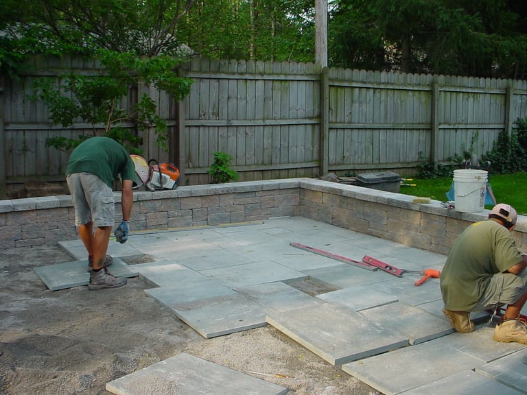 Two workers installing large stone pavers on a residential patio with wooden fence in background