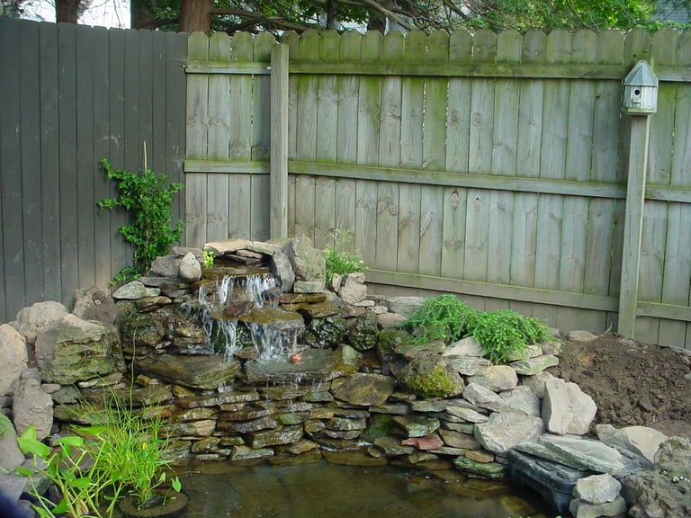 Tiered stone water feature with small waterfall in backyard garden surrounded by moss-covered rocks and wooden fence