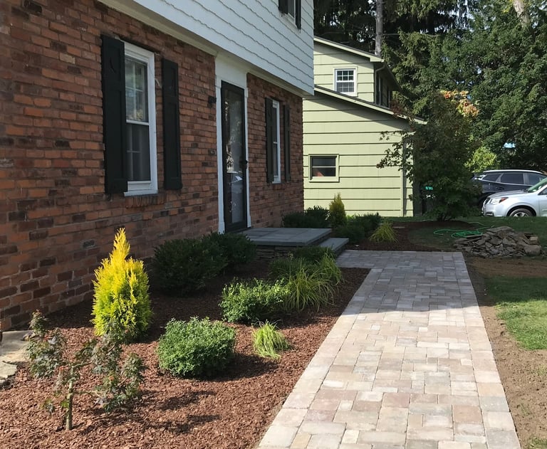 Front entrance of brick house with paver walkway, landscaping, and cream-colored attached garage visible on right