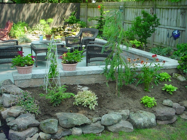 Backyard garden with rock border, planted beds, metal trellis, and patio seating area surrounded by greenery