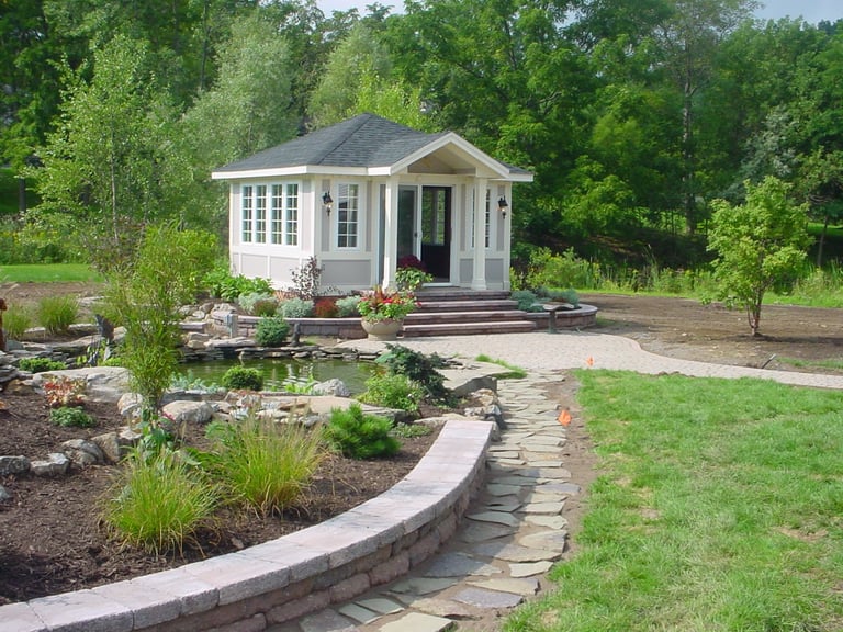 Small modern white cottage with gray roof surrounded by landscaping, stone pathways, and green lawn with trees in background