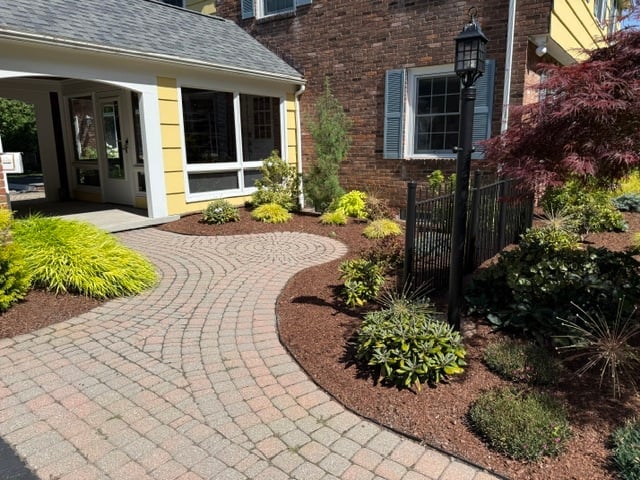 Brick patio with curved pathway leading to modern home entrance, surrounded by landscaped gardens with shrubs and mulch
