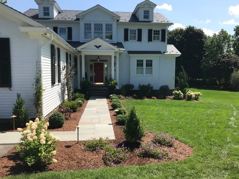 Two-story white colonial house with red door, brick walkway, landscaped garden, and green lawn under blue sky