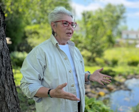 Older man with gray hair and glasses wearing a light blue shirt gestures while standing by a tree near a pond with green landscape behind him