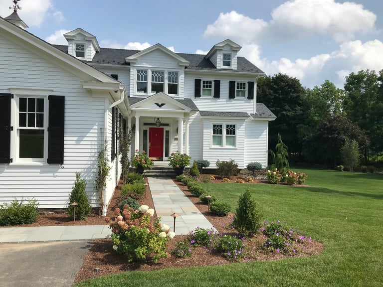 White two-story colonial house with red front door, brick walkway, landscaped gardens, and green lawn under blue sky.
