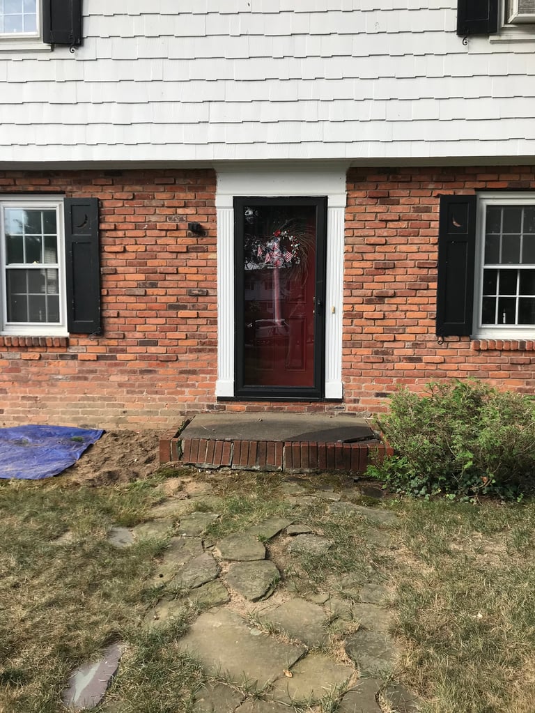 Front facade of a brick house with white trim, dark red door, black shutters, and a front yard with irregular stone pathway and patchy grass