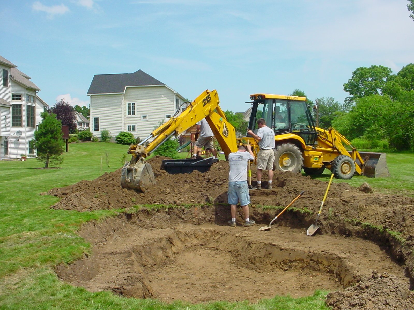 Construction worker with shovel beside yellow excavator digging trench in residential yard near houses