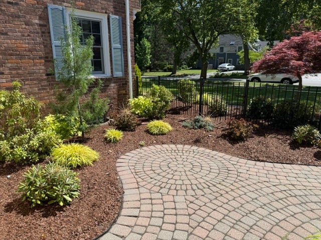 Brick patio with circular paver pattern surrounded by landscaping, shrubs, and a fenced yard near a brick house