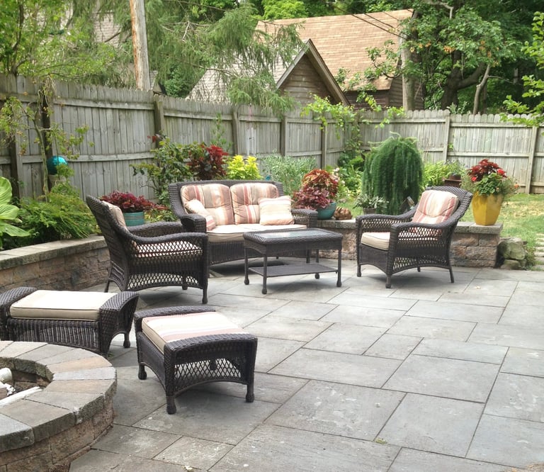 Black wicker patio furniture set with cushions on stone patio, surrounded by flowering plants and wooden fence