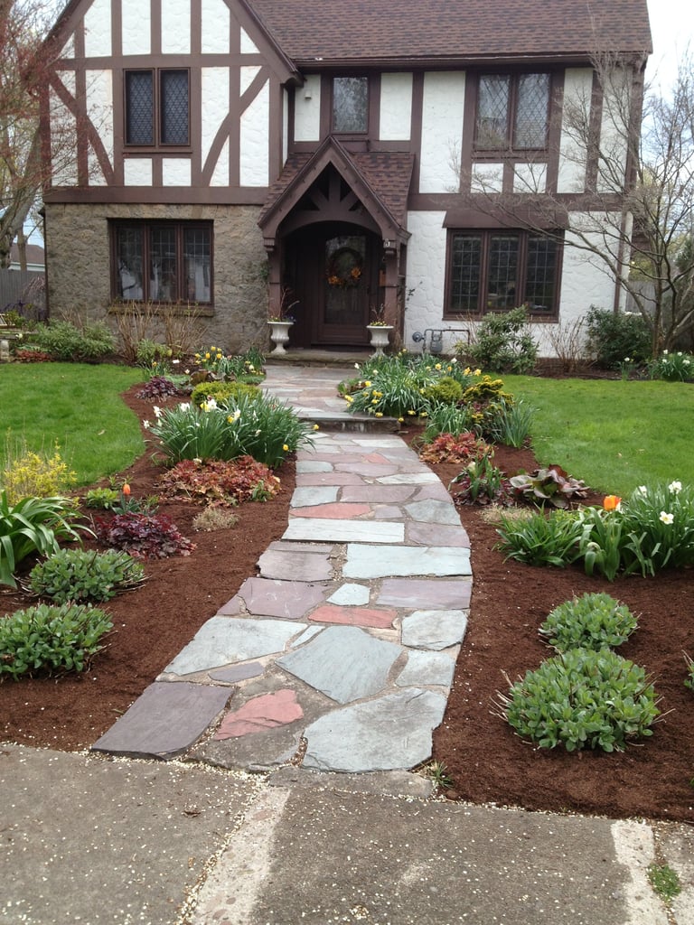 Tudor-style house with stone and brick front, featuring a curved stone walkway through landscaped gardens with flowering plants and shrubs