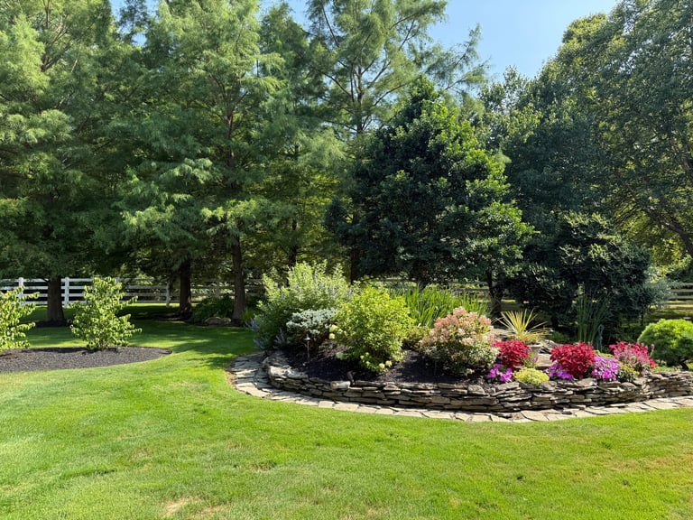 Manicured garden landscape with flowering shrubs, mulched beds, lush green grass, mature trees, and white wooden fence in background