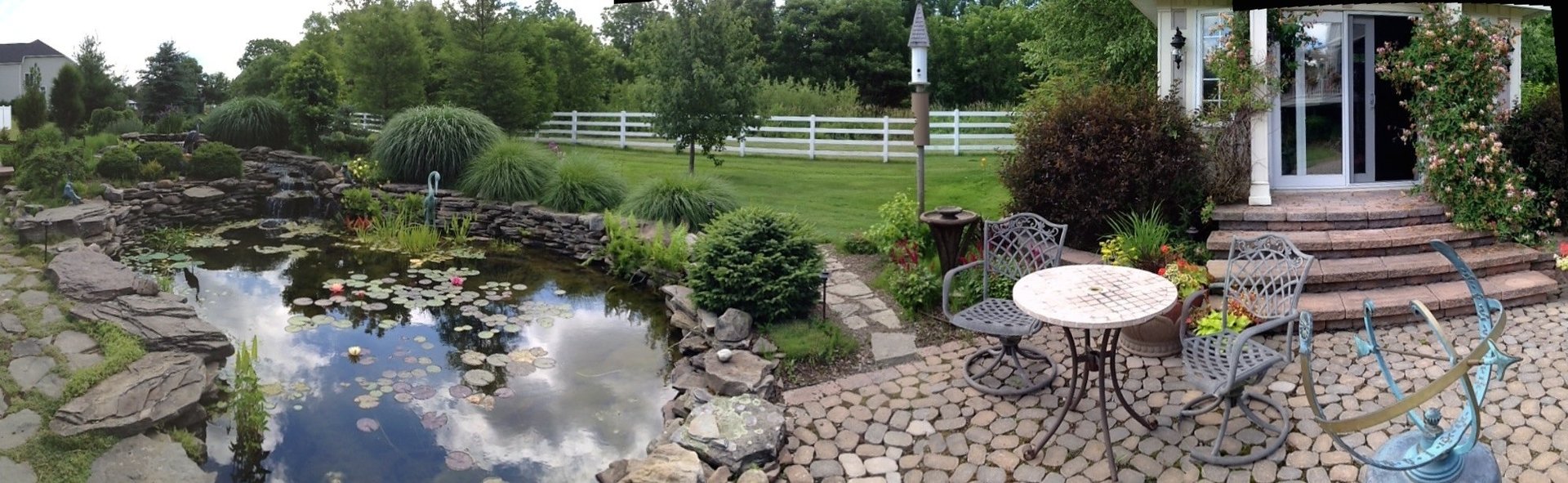 Backyard landscape with pond, patio furniture, brick walkway, and rural property with white fence and trees
