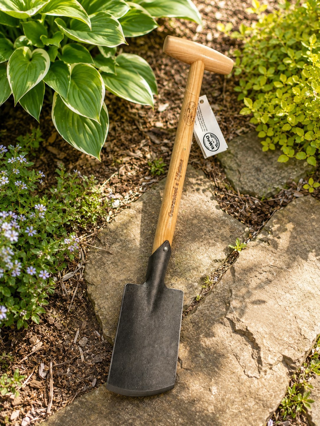 Garden spade with wooden handle and black metal blade resting on gravel surrounded by green plants and foliage