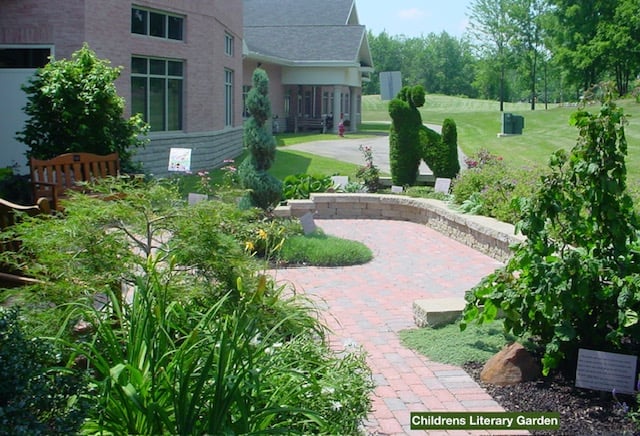 Childrens Library Garden with curved brick pathway, green landscaping, tall shrubs, and residential home in background