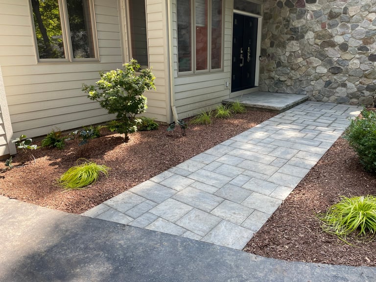 A home entrance with a gray paver walkway leading to a black front door, flanked by mulch landscaping and low shrubs