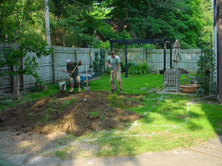 Two people working on a garden bed in a fenced backyard with green grass and wooden fence