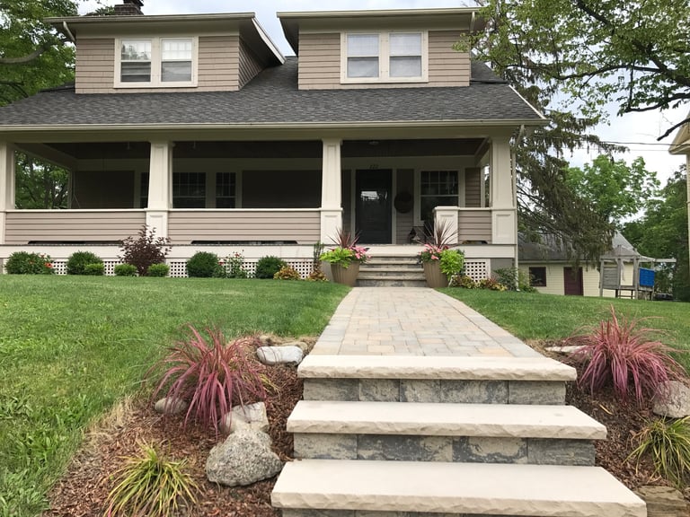 Two-story suburban home with beige siding, front porch, stone walkway and landscaping with ornamental grasses and flower beds