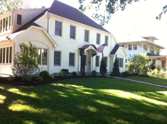 White two-story colonial house with dark roof, front lawn, landscaping, and American flag on entry