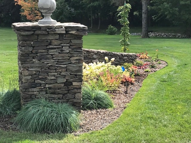 Stone pillar with finial in a garden landscape, surrounded by ornamental grasses and flowering plants along a mulch pathway