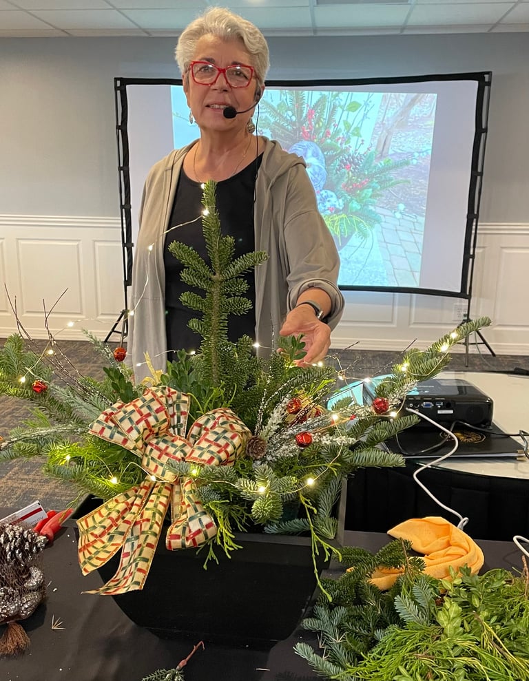 Woman wearing glasses and cardigan standing behind holiday floral arrangement with red ribbon bow in indoor setting